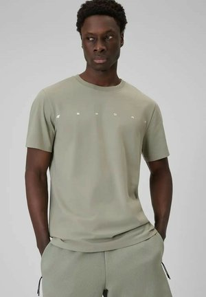 Man die een licht olijfgroene sport-T-shirt en bijpassende korte broek draagt, staat met handen in de zakken tegen een grijze achtergrond.