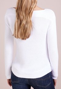 Pull en tricot blanc avec un motif texturé, décolleté large et manches longues, associé à un jean bleu. L'ourlet est légèrement arrondi.