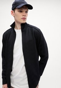 Giacca nera con zip e motivo geometrico testurizzato, abbinata a una t-shirt bianca. Accentuata da un cappellino da baseball blu navy con logo.