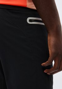 Κοντινή λήψη μαύρης τσέπης με φερμουάρ σε μαύρο παντελόνι, με αντανακλαστικό περίγραμμα, που φοριέται από άτομο με πορτοκαλί μπλούζα, χέρι χαλαρό δίπλα στο σώμα.