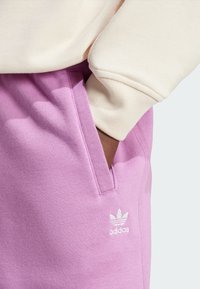 Pantalons de survêtement rose clair avec une texture douce et des poches latérales. Présente un logo Adidas blanc près de la poche et une manche de sweatshirt crème.