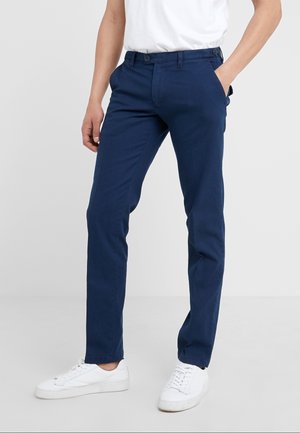 Mężczyzna w granatowych, dopasowanych spodniach, białym pasku i białych trampkach, stojący z jedną ręką w kieszeni.