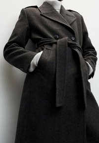 Manteau en laine gris foncé avec une coupe croisée, taille ceinturée, revers à encoches et poches latérales. Tissu texturé et lignes épurées.