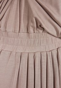 Jasnoróżowa sukienka z marszczoną gumką w talii, plisowaną spódnicą i miękką, gładką fakturą. Prosty krój skupia się na komforcie i układaniu materiału.