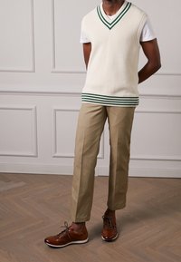 Beiges Strickweste mit grünem Rand, kombiniert mit khakifarbenen Hosen. Braune Schuhe mit Schnürdesign und weißer Sohle. Einfaches, strukturiertes Outfit.