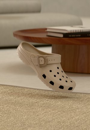 Beige instapklomp met ventilatiegaten en een verstelbare band, geplaatst naast een houten salontafel met gestapelde boeken.