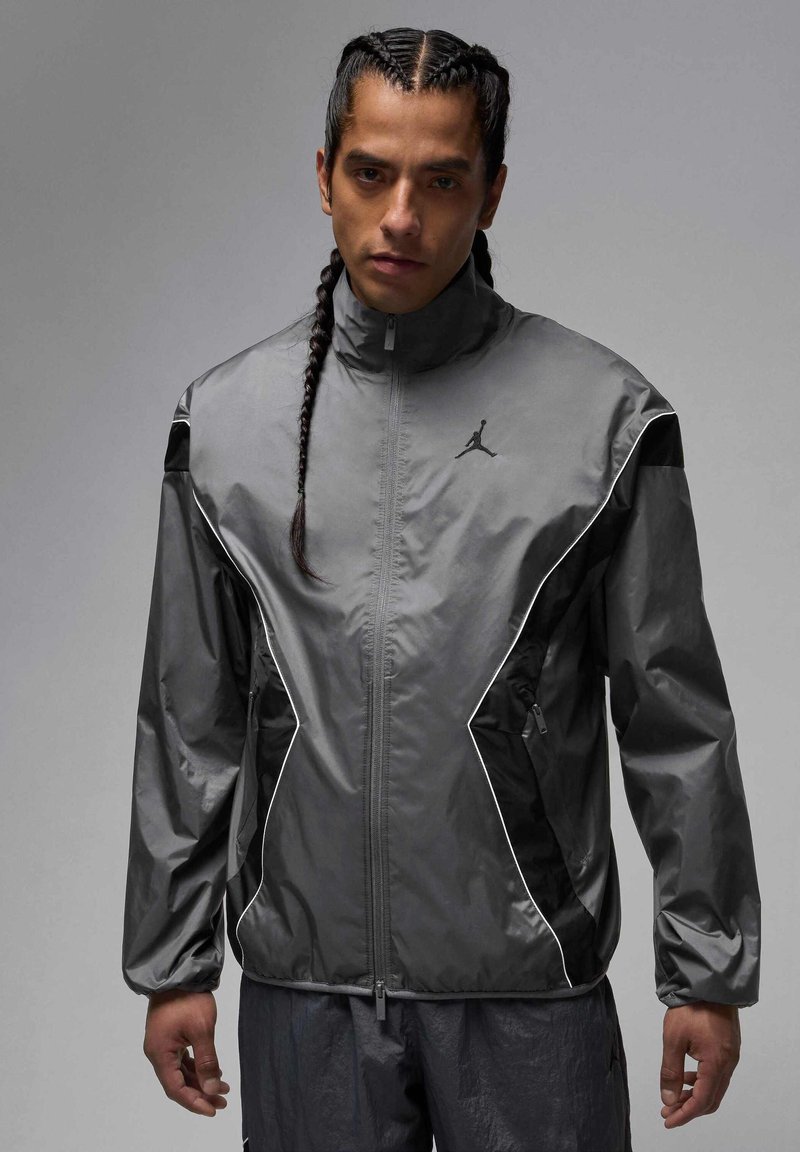 Grå sportjacka med hög krage, dragkedja framtill, svarta accenter och reflekterande piping. Har en logotyp på vänster bröst.