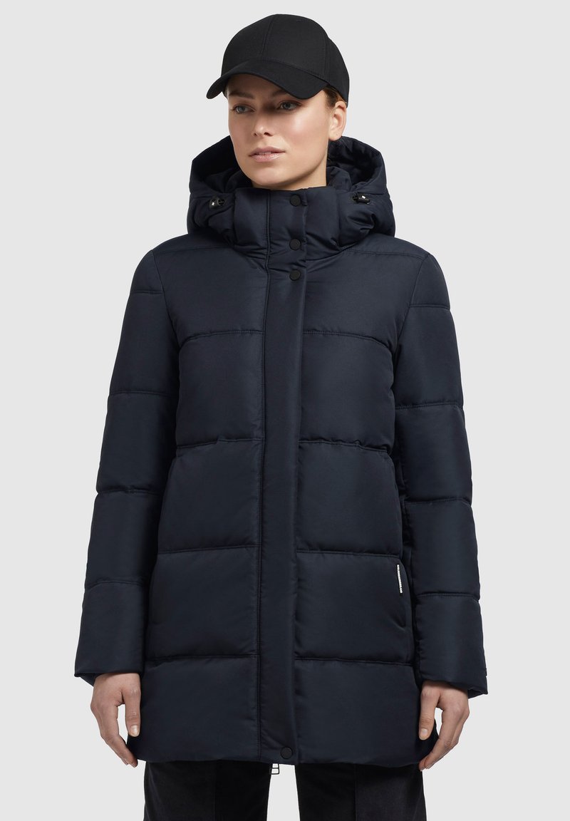 Woman wearing a black padded winter coat with a high collar and hood, paired with a black baseball cap against a plain background.
