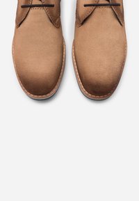 Une paire de chaussures en suède beige à lacets noirs et semelles en cuir cousues, vue de dessus sur un fond blanc.