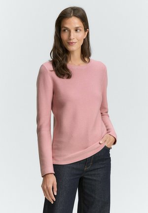 Donna con lunghi capelli castani che indossa una maglietta a maniche lunghe di un rosa chiaro a coste e jeans scuri, in piedi con una mano in tasca.