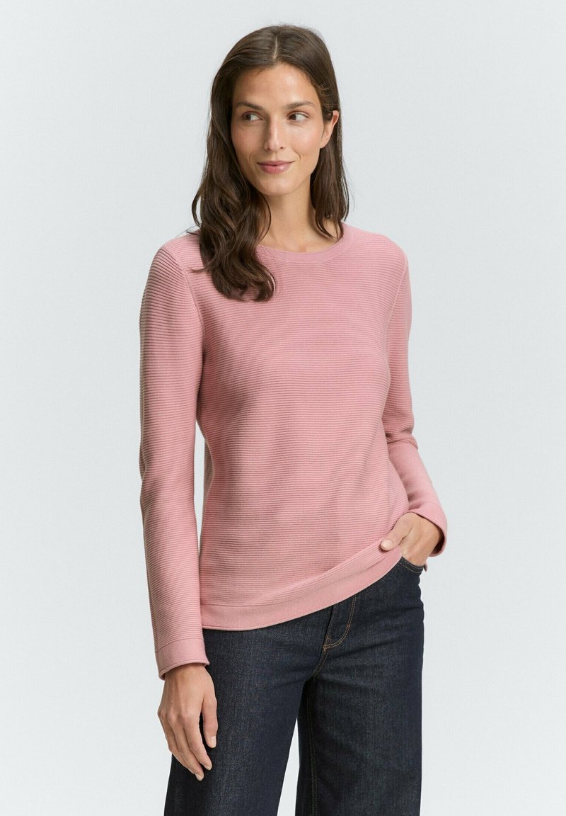 Vrouw met lang bruin haar, gekleed in een lichtroze ribbelshirt met lange mouwen en donkerblauwe spijkerbroeken, staand met een hand in de zak.