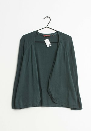 Ciemnozielony kardigan z otwartym przodem i długimi rękawami, wiszący na czarnym wieszaku, z białą metką cenową przymocowaną do kołnierza.