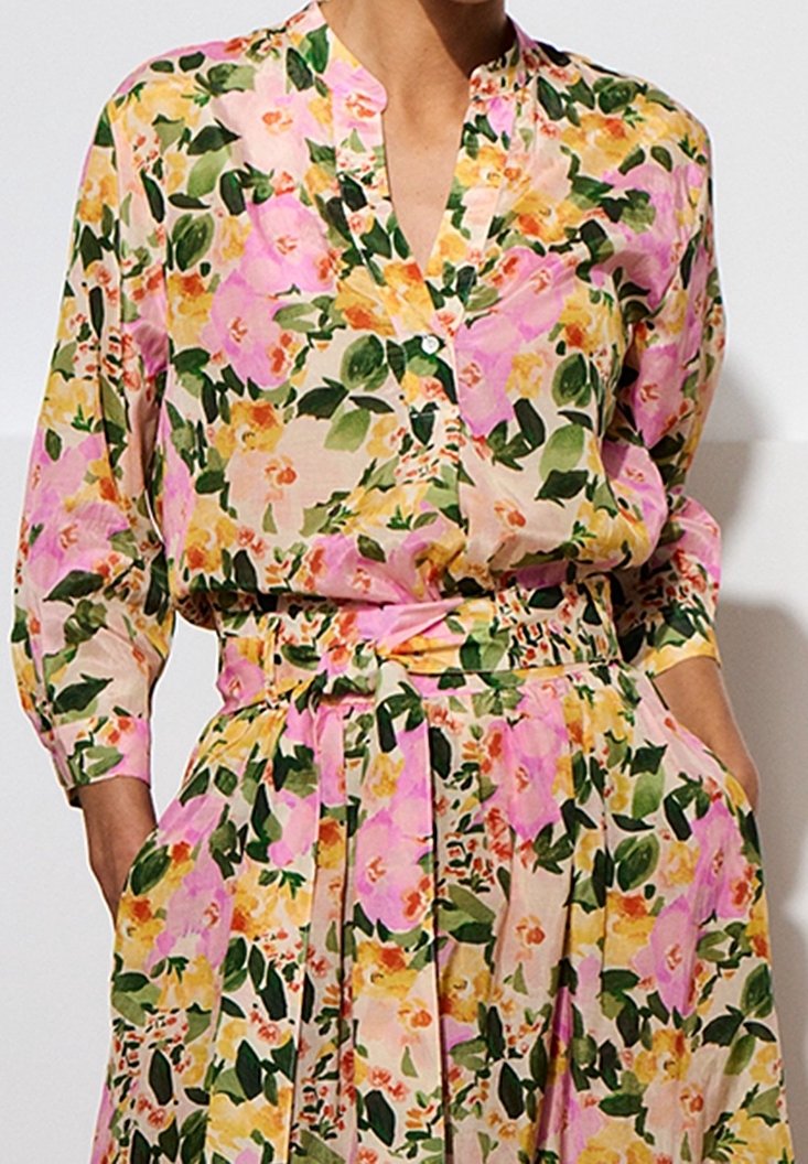 Femme portant une robe fleurie avec des fleurs roses, jaunes et vertes, manches longues, taille ceinturée et mains dans les poches.
