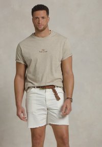 Bežs kokvilnas t-krekls ar mazudrukātu logo, kombinēts ar baltām džinsu šortiem ar saplēstu apakšmalu un brūnu austu jostu.