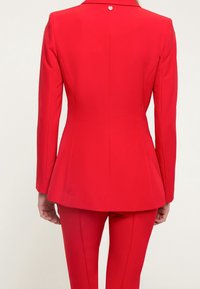 Röd skräddarsydd kavaj med peplumform, hög krage och en knapp i ryggen. Slät textur, tillsammans med figursydda röda byxor.