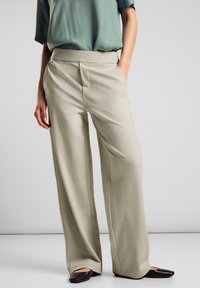 Ljust beige vida byxor i mjukt material med slät front och sidofickor, tillsammans med en löst sittande grön topp och svarta skor.