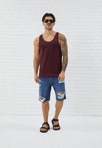 Maroon tank top, shorts denim yang rusak, sandal hitam. Model berpose di depan dinding bata putih. Fitur termasuk tato dan kacamata hitam.