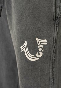 Donkergrijze sweatpants met een wit grafisch logo op de dij, met een zachte textuur en zichtbare stikseldetails.