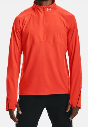 Homme portant un pull-over de sport à manches longues orange vif avec une fermeture éclair au quart et un logo blanc Under Armour sur le col, ainsi qu'un pantalon de sport noir.