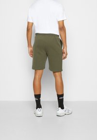 Pantaloni corti verde oliva con vita elastica, lunghezza fino a metà coscia. Calze bianche e scarpe da ginnastica completano il look sportivo. È visibile una maglietta bianca.