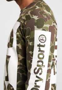 Camisa de manga larga con estampado de camuflaje en tonos verdes, con detalles en las mangas en blanco que presentan el texto "DrySport" y acentos del logo.