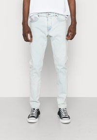 Jean en denim bleu clair avec une coupe droite, un design à cinq poches et une fermeture à boutons sur le devant. Porté avec des baskets noires et blanches.