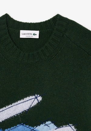 Grønn strikket genser med ribbet rund hals og et blått og hvitt broderi med kontrasterende sømdetaljer.