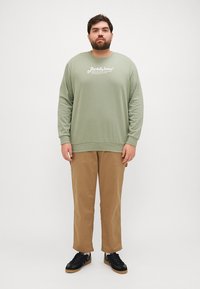 Groene sweatshirt met wit logo, gedragen met kaki broek en zwarte sneakers. De stof lijkt zacht; de sweatshirt heeft een ontspannen pasvorm en lange mouwen.