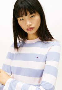 Camisa de manga larga a rayas en rosa claro y azul, con cuello redondo y un pequeño logo en el pecho. Textura suave y lisa.