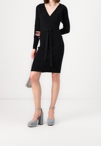 Abito nero a coste con scollo a V, maniche lunghe e cintura abbinata. Tacchi platform in suede grigio con cinturino alla caviglia e tacco stondato.
