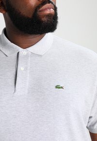 Polo gris hecho de tejido texturizado, con cuello y tapeta de dos botones, y un pequeño logo de cocodrilo verde en el pecho.