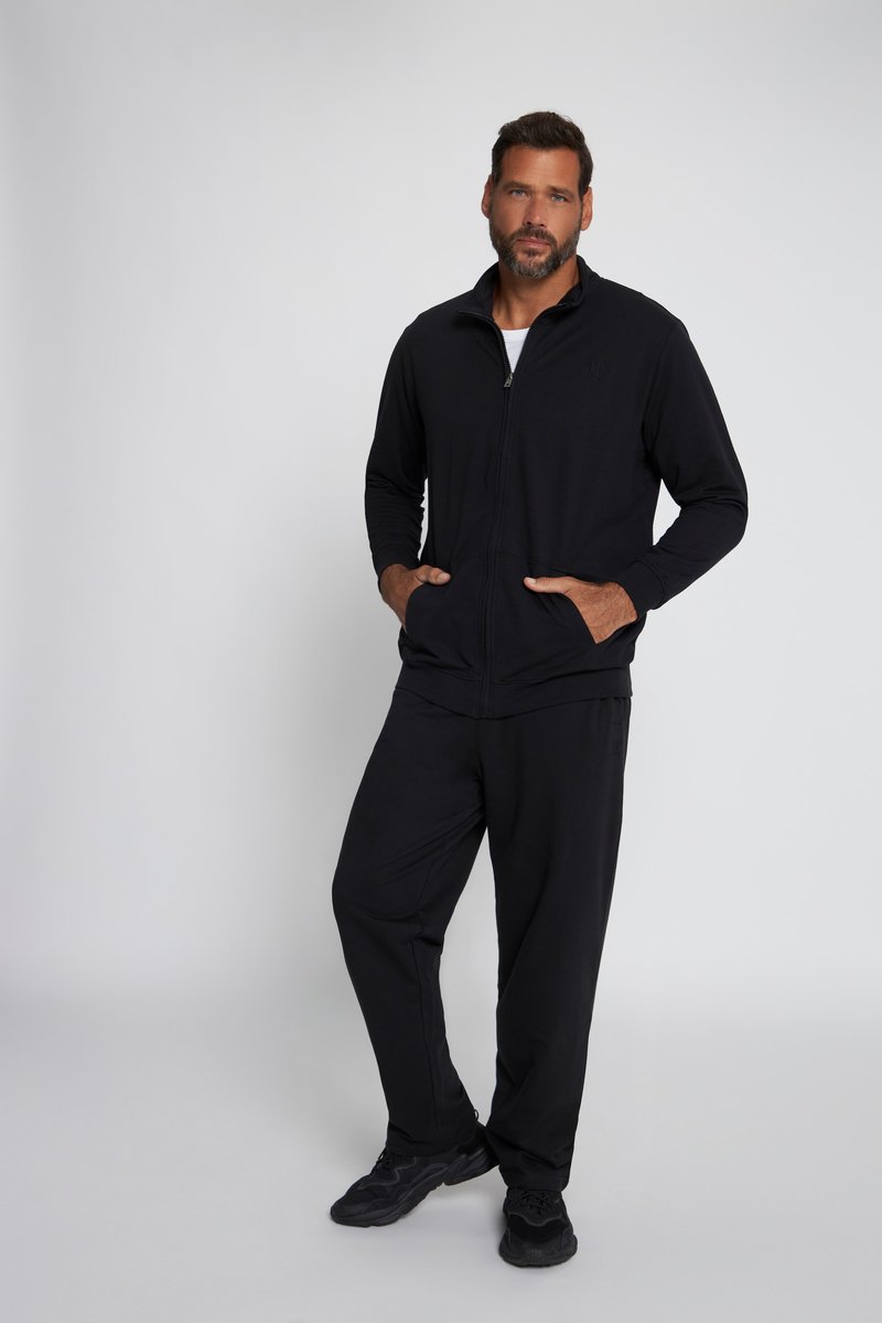 Homme barbu portant une veste zippée noire, un pantalon noir et des baskets noires, debout les mains dans les poches devant un fond gris.