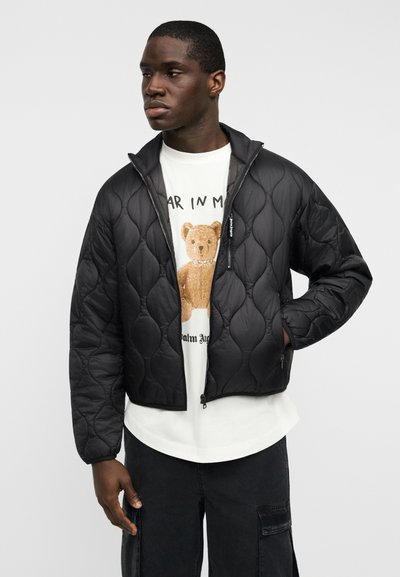 Μαύρο μπουφάν με καπιτονέ σχέδιο, όρθιο γιακά, κλείσιμο με φερμουάρ και πλαϊνές τσέπες. Φορεμένο πάνω από λευκό t-shirt με σχέδιο και μαύρο παντελόνι cargo.