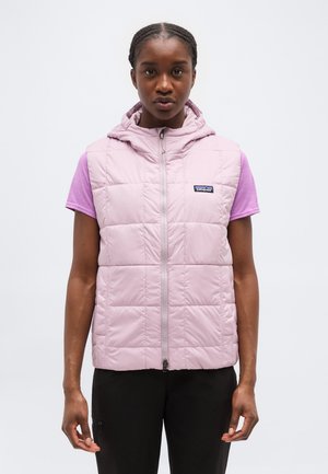 Junge Frau mit geflochtenem Haar, die eine hellrosa gesteppte ärmellose Kapuzenweste über einem lila kurzärmeligen Shirt und schwarzen Hosen trägt.