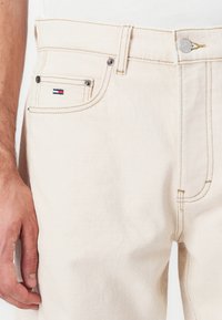 Vaqueros de mezclilla beige con corte recto, costuras visibles, bolsillos laterales y un pequeño logo en rojo, blanco y azul en el bolsillo frontal.