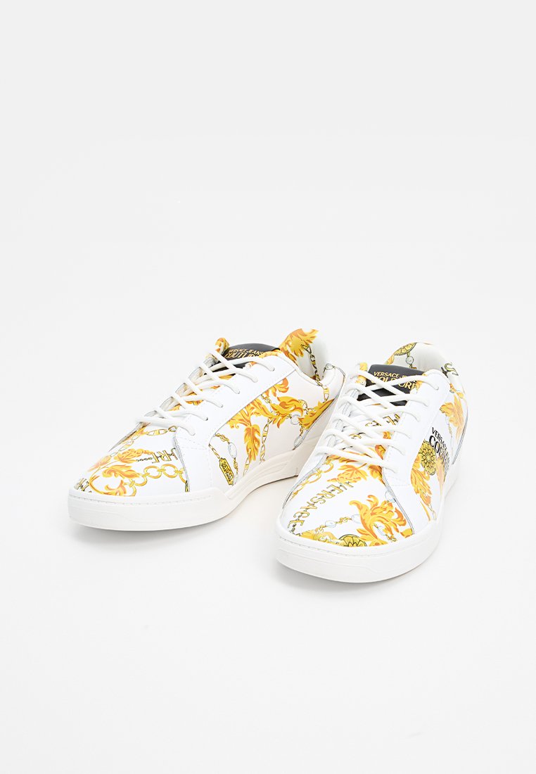 Baskets blanches avec un motif orné jaune vif et doré, dotées d'un système de laçage et de semelles en caoutchouc. Les motifs comprennent des chaînes et des accents floraux.