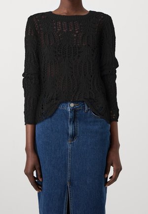 Kvinde iført en sort hæklede langærmet top kombineret med et højttaljet blåt denimskørt med frontslids og knaplukning.