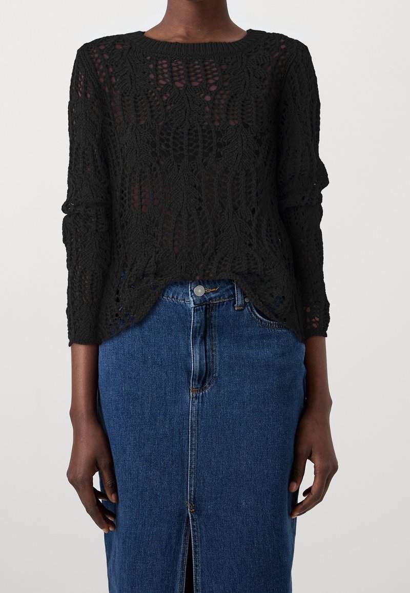 Femme portant un haut noir à manches longues en crochet associé à une jupe en jean bleue taille haute avec une fente avant et une fermeture à boutons.