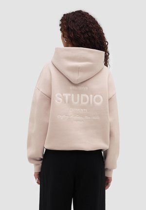 Žena s kudrnatými vlasy nosí béžovou mikinu s kapucí s nápisem "WRSTBHVR STUDIO GARMENTS" na zádech, v kombinaci s černými kalhotami, otočená zády.