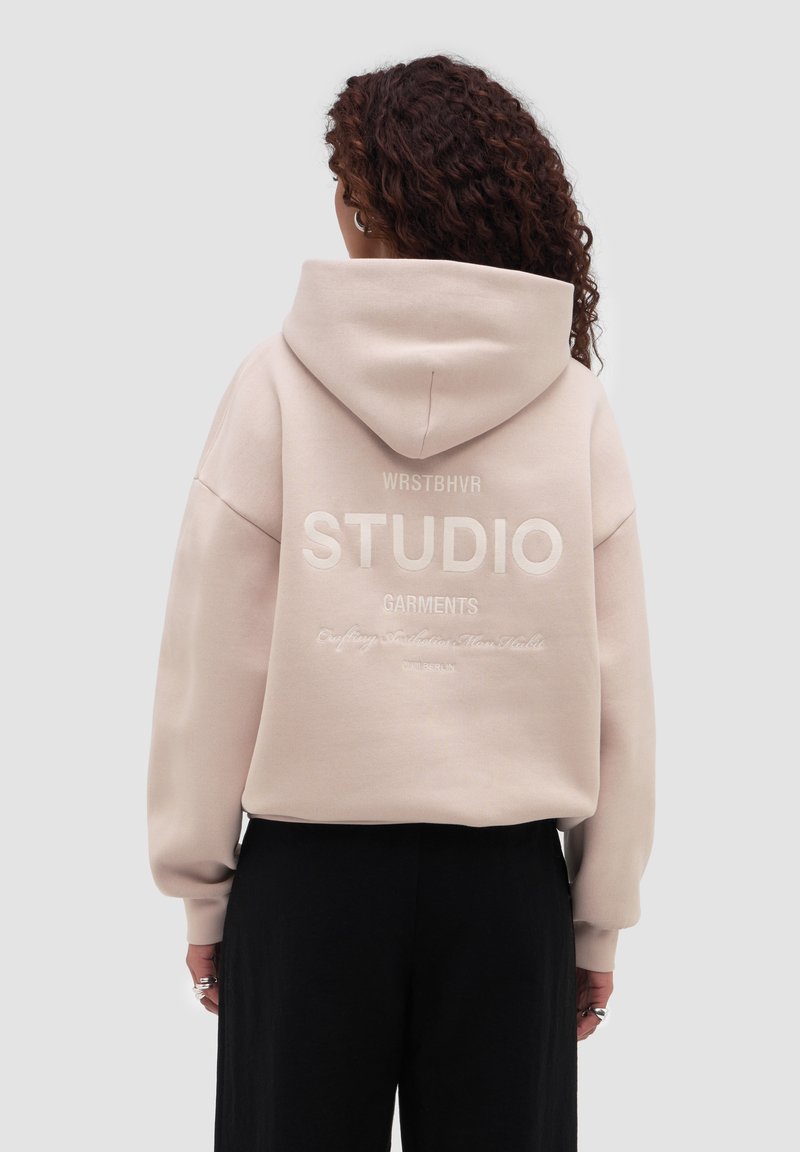 Vrouw met krullend haar draagt een beige hoodie met de tekst "WRSTBHVR STUDIO GARMENTS" op de achterkant, gecombineerd met een zwarte broek, gezicht van de camera afgewend.