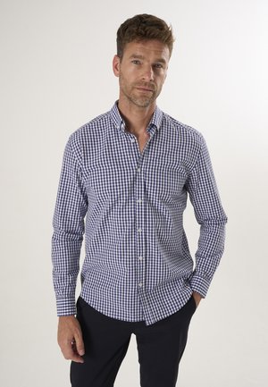 Mann mit kurzen braunen Haaren, der ein blau-weiß kariertes Hemd mit Knöpfen und schwarze Hose trägt, steht mit einer Hand in der Tasche.