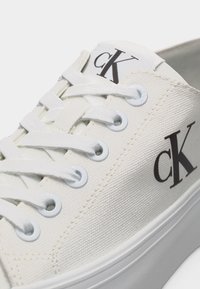 Άσπρο καμβά παπούτσι τύπου σνίκερ με λευκά κορδόνια, μεταλλικές οπές και μαύρο λογότυπο "cK" στη γλώσσα και στη πλευρά κοντά στη σόλα.