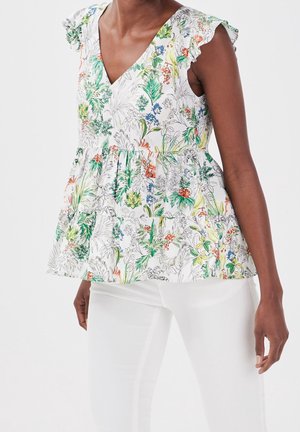 Femme portant un haut sans manches à imprimé floral avec des épaules volantées et un pantalon blanc sur un fond clair uni.