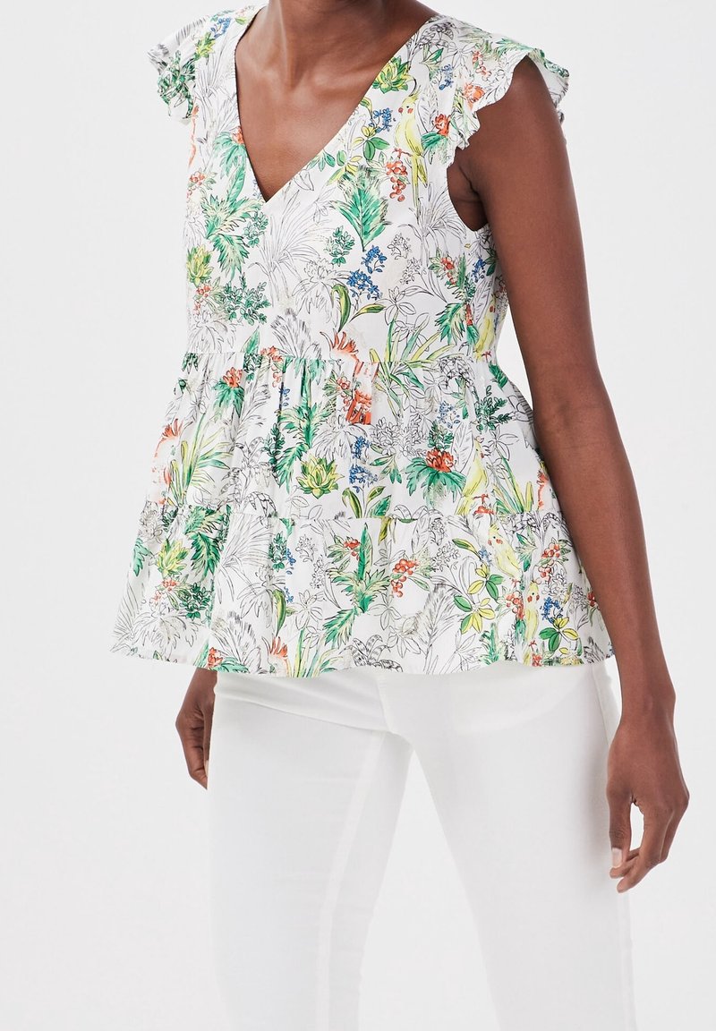 Femme portant un haut sans manches à imprimé floral avec des épaules volantées et un pantalon blanc sur un fond clair uni.