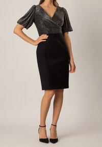 Femme portant une robe noire jusqu'aux genoux avec un haut en V scintillant et des manches bouffantes, associée à des escarpins noirs à bride cheville.