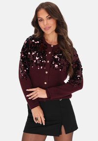 Donna con lunghi capelli castani che indossa un cardigan di paillettes bordeaux con bottoni dorati e una mini gonna nera con uno spacco frontale.