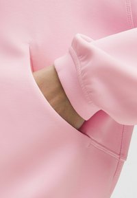 Růžová mikina s hladkou texturou, boční kapsou a elastickými manžetami. Švy jsou viditelné a doplňují design.