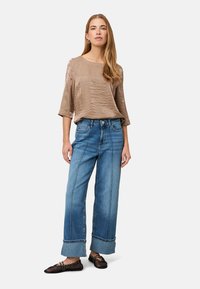 Braun strukturiertes Oberteil mit weiten Ärmeln, kombiniert mit lockeren blauen Jeans aus Denim mit auffälligen Nähten und umgeschlagenen Säumen, getragen mit schwarzen Ballerinas.