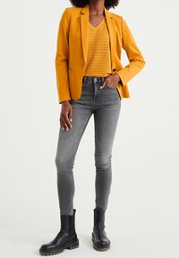 Gul kavaj över en strukturerad gul v-ringad tröja, grå slim fit jeans och svarta ankelskor med tjock sula och rund tå.