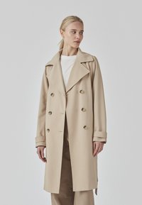 Beige trench coat with a double-breasted design, large lapels, and button accents. Features wide cuffs and straight hemline. Smooth texture.